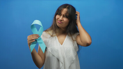 Hispanic woman holding teal awareness ribbon with eyes closed in blue studio; health awareness concern.