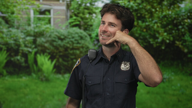 Police officer man making phone hand gesture with radio microphone on uniform while standing by building window; friendly duty.