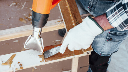 Carpenter using heat gun and wood scraper removes paint from old wooden window. Construction industry, carpentry. Restoration. 