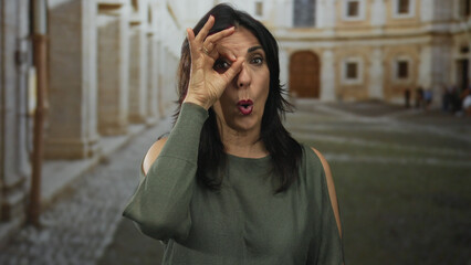 Fototapeta premium Hispanic middle age woman holds hand to eye in ok sign gesture at university building outdoors; approval.