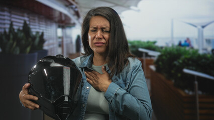 Woman middle age hispanic brunette holds motorbike helmet and presses hand to chest for heartattack at airport; health emergency anxiety.