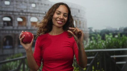 Obraz premium Woman smiling holds a red apple in one hand and pinches fingers with the other on a balcony in front of an ancient building; joy.