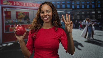 Woman holding a red apple in her left hand and showing four fingers with her right hand in front of a hot dog van on a busy street; health choice happiness.