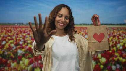 Young woman waving hand and holding a paper gift bag with red heart in a tulip field under blue...