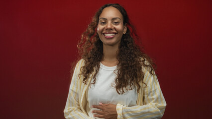 Fototapeta premium Young african american woman with curly hair smiling with hand on stomach and closed eyes wearing a striped shirt in a red studio; joy.