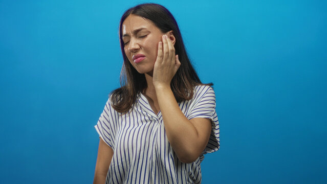 Young hispanic woman touching her ear with hand in blue studio wearing a striped top and closing eyes in pain; discomfort earache.