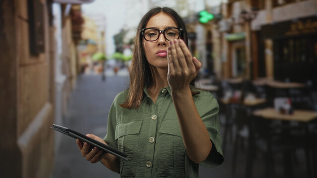 Woman holding tablet beckons with fingers toward viewer at street cafe terrace outdoors wearing glasses; impatience.