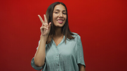 Fototapeta premium Hispanic young brunette woman smiling and showing a peace sign with her hand and visible forearm in a red studio setting; playful.