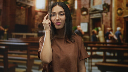 Young brunette woman points finger to temple while standing among wooden pews in a church, facing camera; contemplation faith.