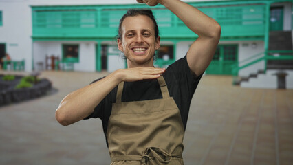 Man smiling with hands framing face at building entrance wearing apron and showing forearms;...