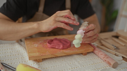 Man shaping clay with paint covered hands on a wooden board in art studio surrounded by small paint pots and sculpting tools; concentration.