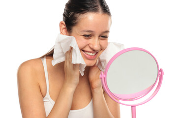 Young smiling woman with long dark hair cleaning her face with wipes in front of a mirror, studio...