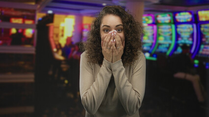 Obraz premium Young woman with hands covering mouth at a casino slot machine smiling with surprised expression; surprise luck celebration.
