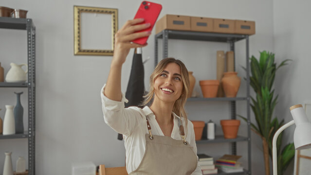 Young smiling blonde woman wearing apron holds smartphone up with extended arm in pottery studio; artistic pride creative passion.