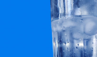 Close-up of a chilled glass of water with condensation and ice cubes against a bright blue background. Symbolizing freshness, hydration, purity and cool refreshment during healthy living.
