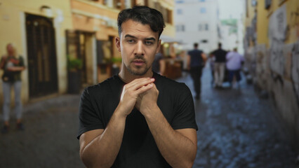 Young hispanic man appearing thoughtful on a busy city street in an outdoor urban setting, surrounded by blurred pedestrians and rustic buildings.