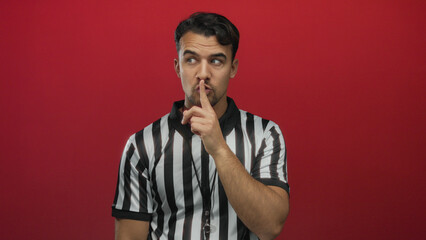 Young hispanic man referee in striped shirt making hush gesture on isolated red background conveying silence and authority.