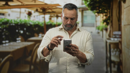 Man with grey beard taps smartphone screen on outdoor restaurant terrace while looking down at device and holding phone with both hands; concentration.