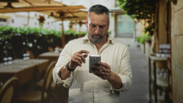 Man with grey beard taps smartphone screen on outdoor restaurant terrace while looking down at device and holding phone with both hands; concentration. - Powered by Adobe