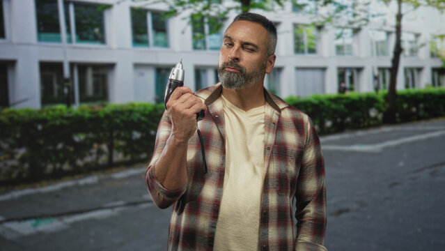 Man holding power drill in right hand on street, carpenter inspecting the tool with raised eyebrow and neutral pose; confidence experience. - Powered by Adobe