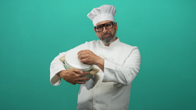 Middle aged hispanic man chef holding a pot and tightening its lid in a teal studio wearing white uniform and hat; culinary focus.