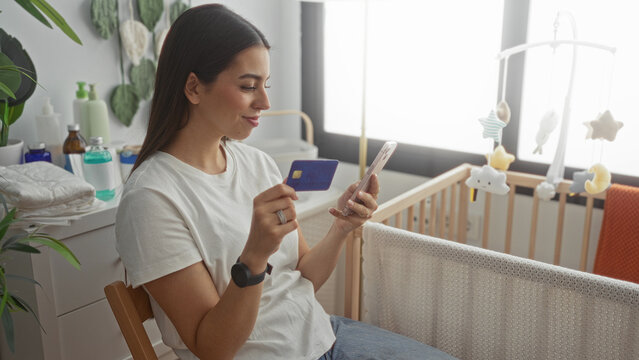 Woman holding creditcard and smartphone in bedroom beside wooden cradle and baby mobile, seated on chair by crib; calm.