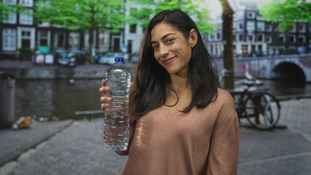 Woman holding a plastic bottle on a street canal in amsterdam, smiling and wearing a v neck sweater; hydration contentment.
