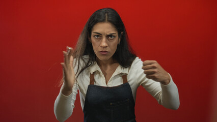 Woman with clenched hands and stern expression making a forward strike gesture in a red studio;...