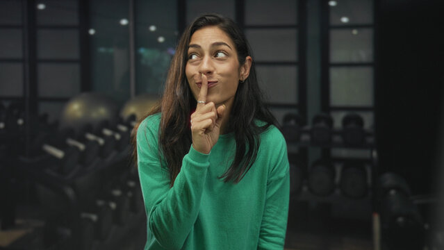Woman finger to lips for silence in gym with dumbbells and kettlebells visible wearing green sweater facing camera; secret silence. - Powered by Adobe