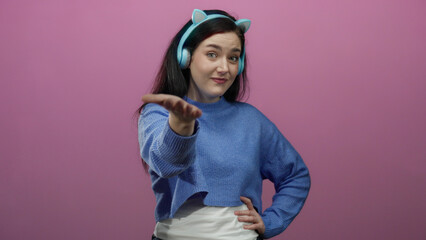 Woman reaching hand to camera wearing headphones and blue sweater against pink background exuding joy and friendliness