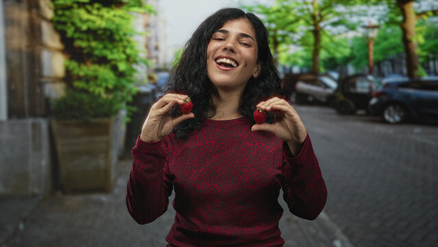 Woman holding strawberries with both hands on a city street wearing a red sweater and smiling broadly; joy playful fun laugh. - Powered by Adobe