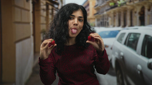 Young arab woman holding strawberries and sticking tongue out on a busy city street; playful summer freshness.