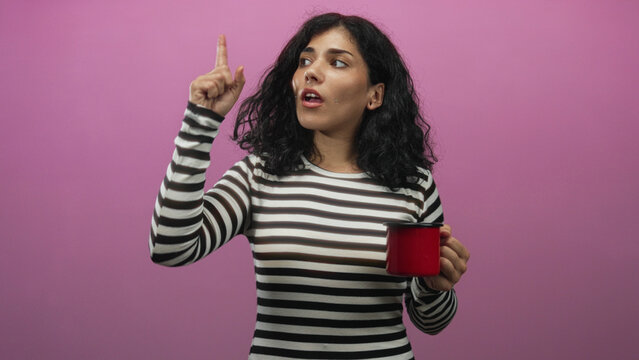 Young woman with brunette hair wearing striped top points finger up while holding red mug in pink studio; thoughtful idea.