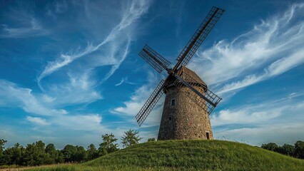 Fototapeta premium A historic windmill on a grassy hill under a bright sky with clouds.
