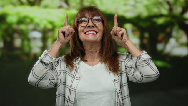 Senior hispanic woman smiling in a vibrant outdoor park setting, pointing upwards with both fingers, conveying happiness and positivity amidst a lush green backdrop