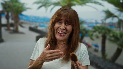 Hispanic senior woman enjoying a cookie at the seaside with palm trees in the beach background.