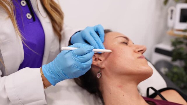 Professional beautician in blue gloves drawing guidelines on a woman's face before a cosmetic procedure in a modern beauty clinic