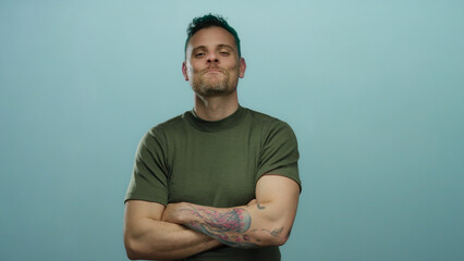 Young man with tattoos and crossed arms stands confidently against a blue background, wearing a green shirt, smiling with a modern hairstyle, showcasing a stylish and attractive expression.