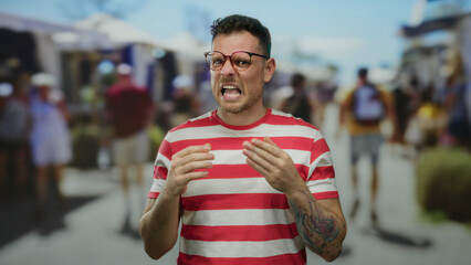 Naklejka premium Man in striped shirt with tattoos makes expressive gestures on a busy urban street filled with people and vibrant activity under a clear blue sky.