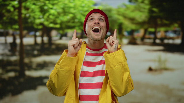 Hispanic man smiling joyfully outdoors in a park, wearing a red beanie and yellow raincoat, with lush green trees in the background.