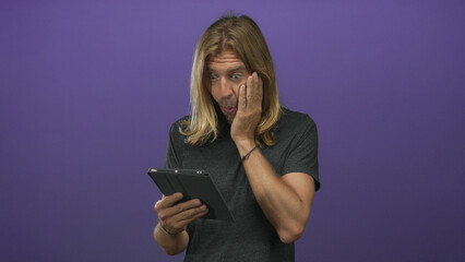 Blond man with long hair in dark shirt points finger to tablet with mouth open in purple studio;...