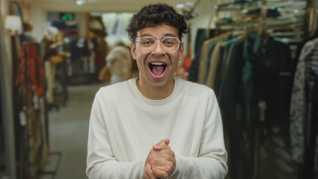 Young hispanic man with glasses clasping hands laughing in a retail building aisle under warm lights; joy.