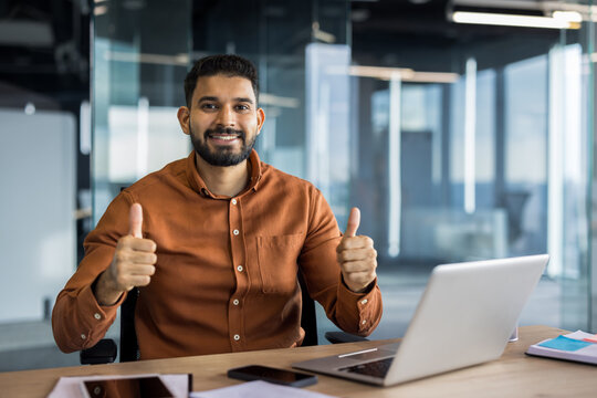 Indian businessman sitting at a modern office desk with a laptop, smiling confidently and giving a dual thumbs up, symbolizing approval, positivity, and achievement in his professional career