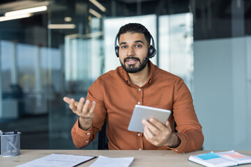 Young man wearing a headset with a microphone sitting at a desk, looking at the camera and gesturing while holding a digital tablet, representing online support and communication
