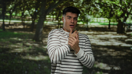 Young man in striped shirt appears worried, covering mouth in a sunlit park surrounded by lush trees suggesting a contemplative mood in serene outdoor setting.