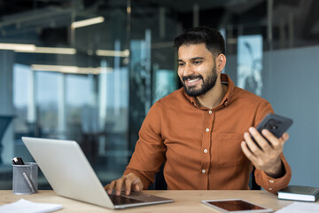 Smiling young man with a beard sitting at a wooden desk in a contemporary office, multitasking by typing on a laptop and holding a smartphone, representing connectivity and productivity