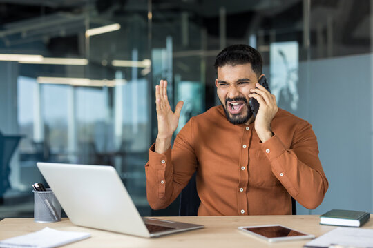Businessman in modern office yelling into mobile phone while angrily gesturing at laptop, stressed and frustrated during a heated work call and urgent conflict at his desk