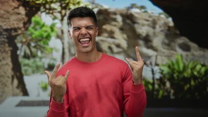 Young man making fun gestures on a city street in a red sweater outdoors with trees in the background, smiling energetically during a sunny day.