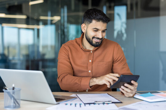 Young bearded man smiling, working with a calculator, documents, and laptop on a desk in a modern office, representing finance management and business success