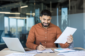 Young smiling businessman at his modern office desk signing documents, surrounded by laptop, charts and paperwork as he reviews financial data and plans strategy
